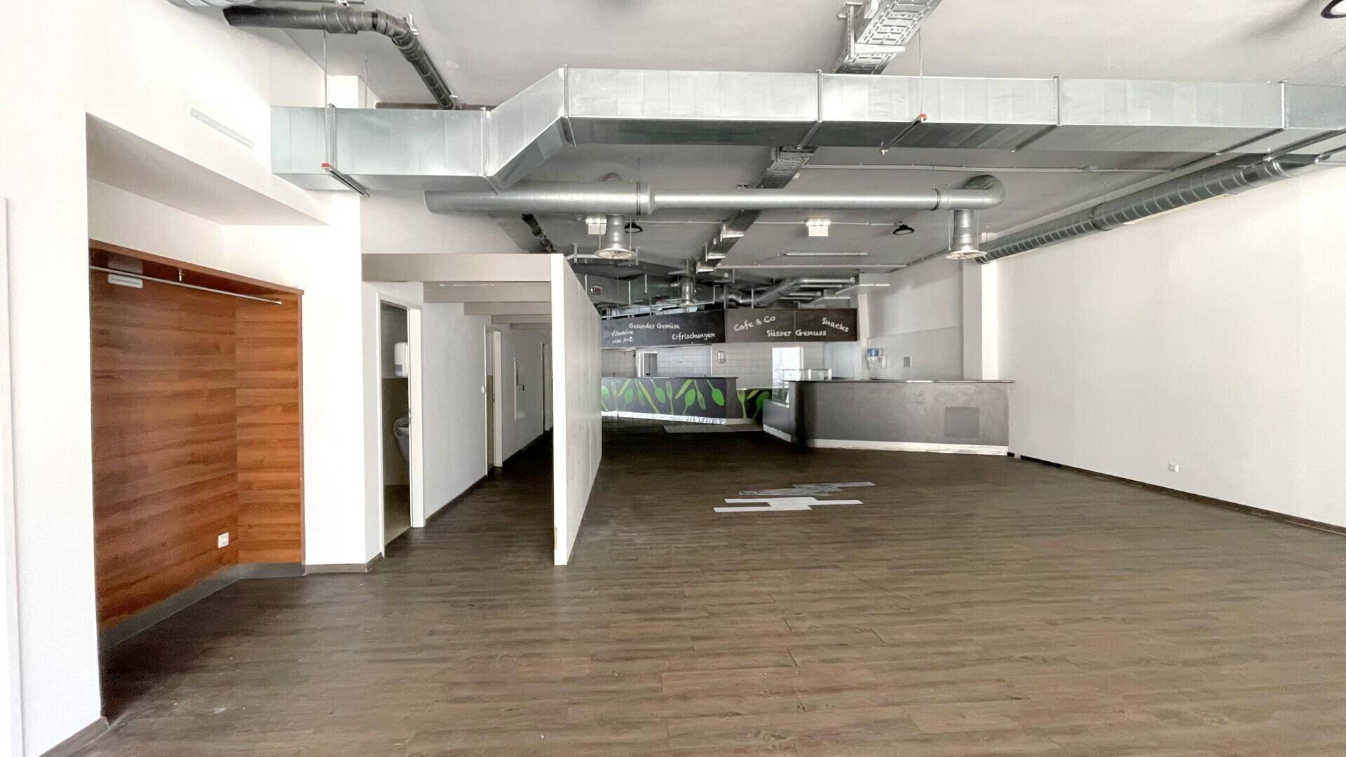 Geräumiger Gewerberaum mit offener Deckenkonstruktion, sichtbaren Lüftungsrohren und einem Thekenbereich im Hintergrund.