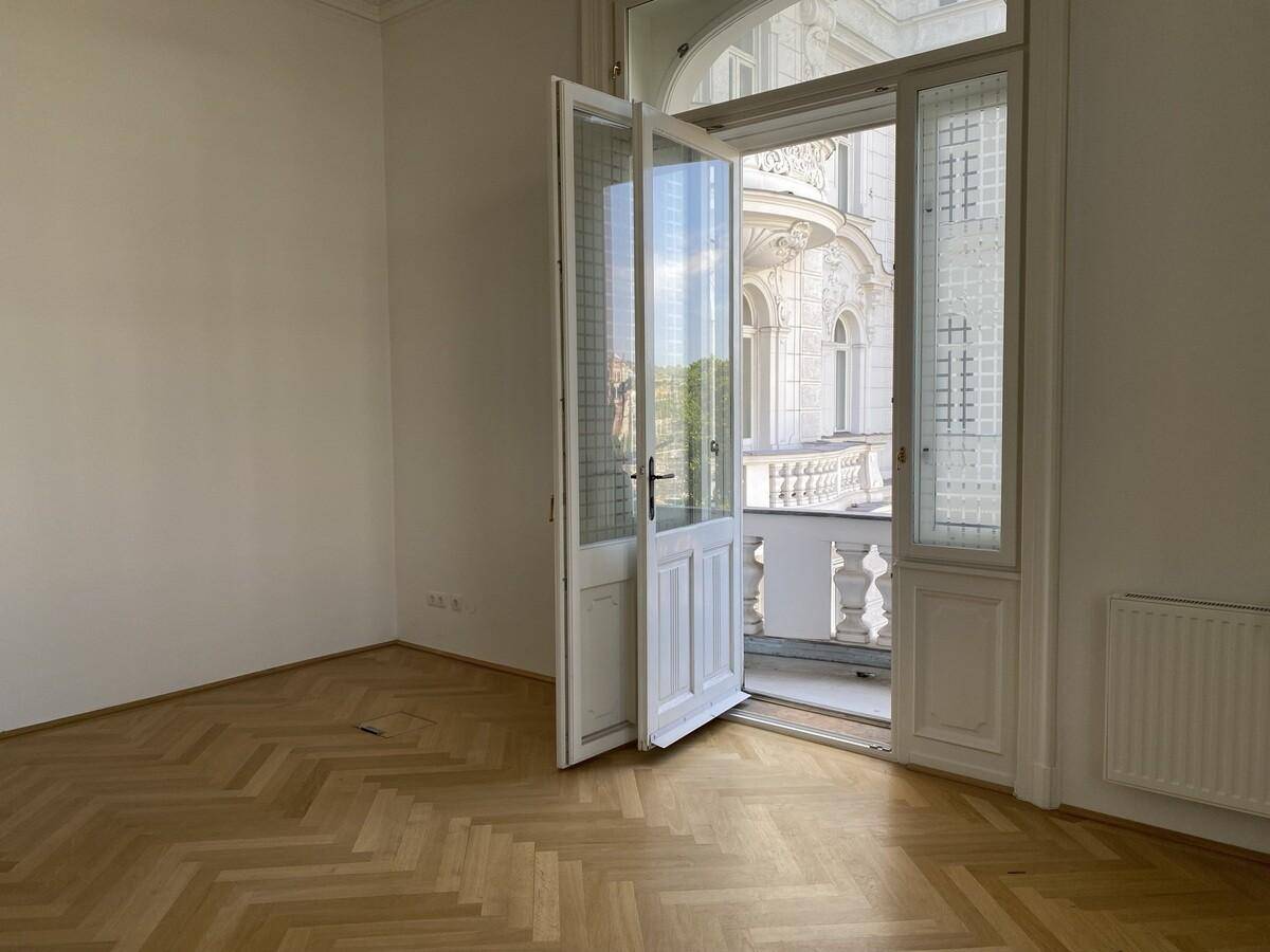 Heller Raum mit Fischgrätparkett und einer geöffneten Fenstertür, die Zugang zu einem Balkon bietet.