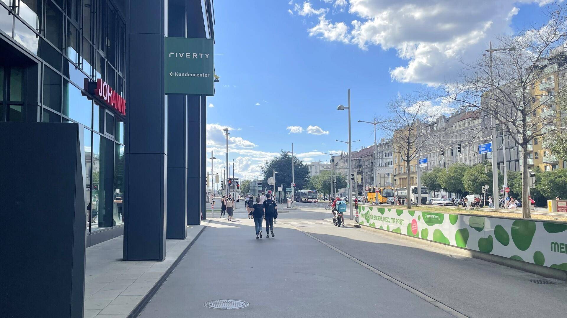 Weitläufige Straßenansicht mit modernen Gebäuden, Fußgängern und blauem Himmel an einem sonnigen Tag.