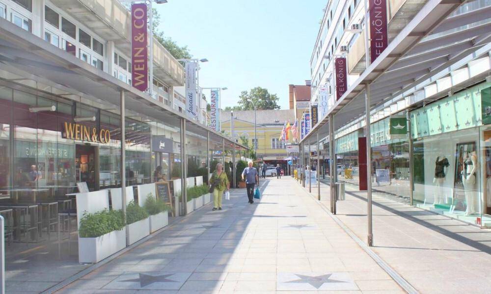 Belebte Einkaufsstraße mit Geschäften, Restaurants und Fußgängern an einem sonnigen Tag in der Stadt.