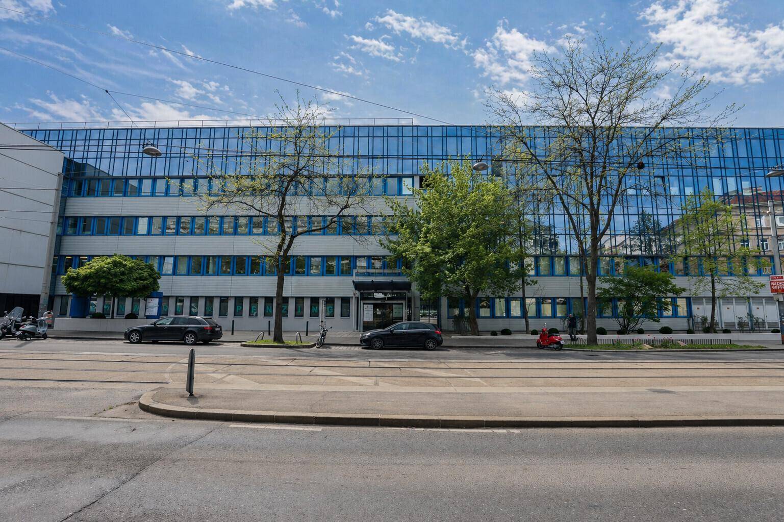 Ansicht der langen Fassade eines modernen Bürogebäudes mit blauen Fenstern und Bäumen an einer Straße.