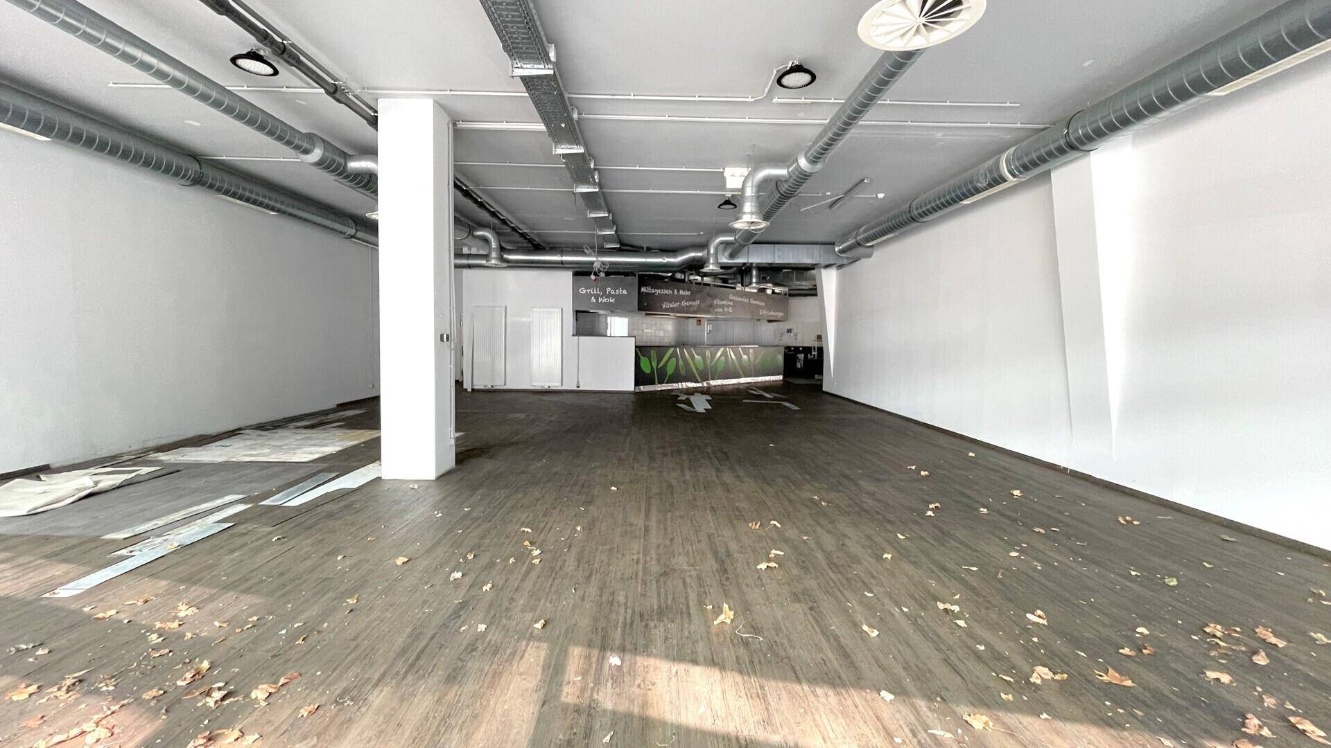 Weitläufiger, leerer Gewerberaum mit Säulen, sichtbaren Deckeninstallationen und Laub auf dem Boden.