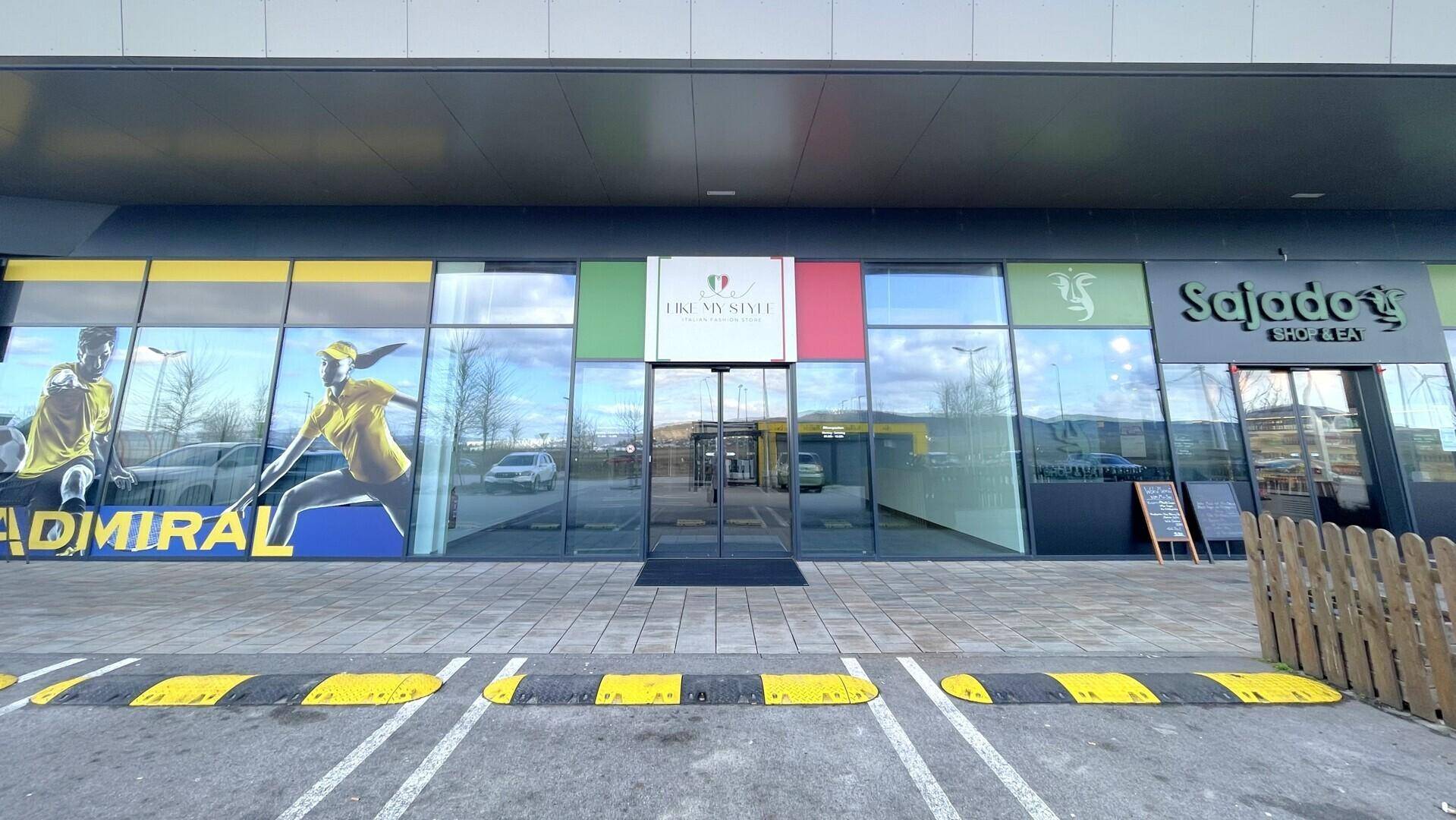 Moderne Ladenfronten eines Einkaufszentrums mit großen Glasfenstern und farbigen Werbeflächen entlang des Parkplatzes.