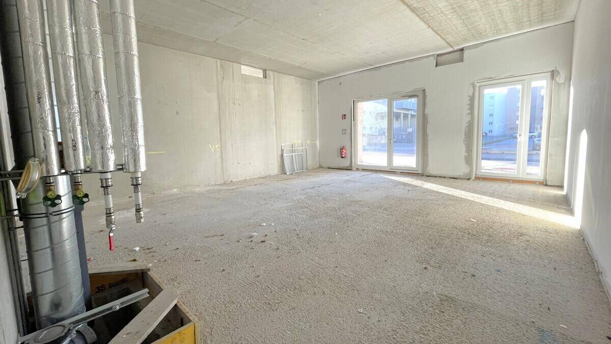 Unfertiger Gewerberaum mit Betonwänden, großen Fenstern und sichtbaren Rohren an der Decke.