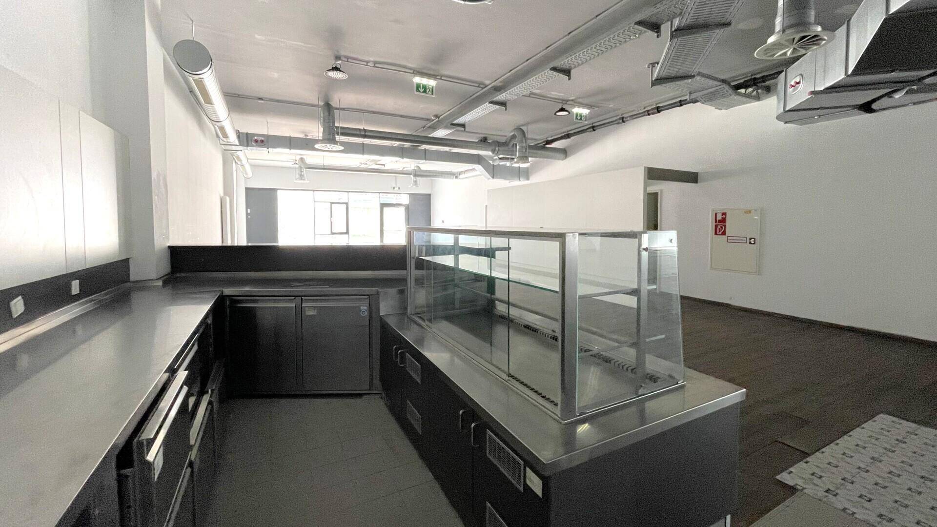 Gewerbliche Küche mit Edelstahlarbeitsflächen, Kühlvitrine und sichtbaren Lüftungsrohren an der Decke.