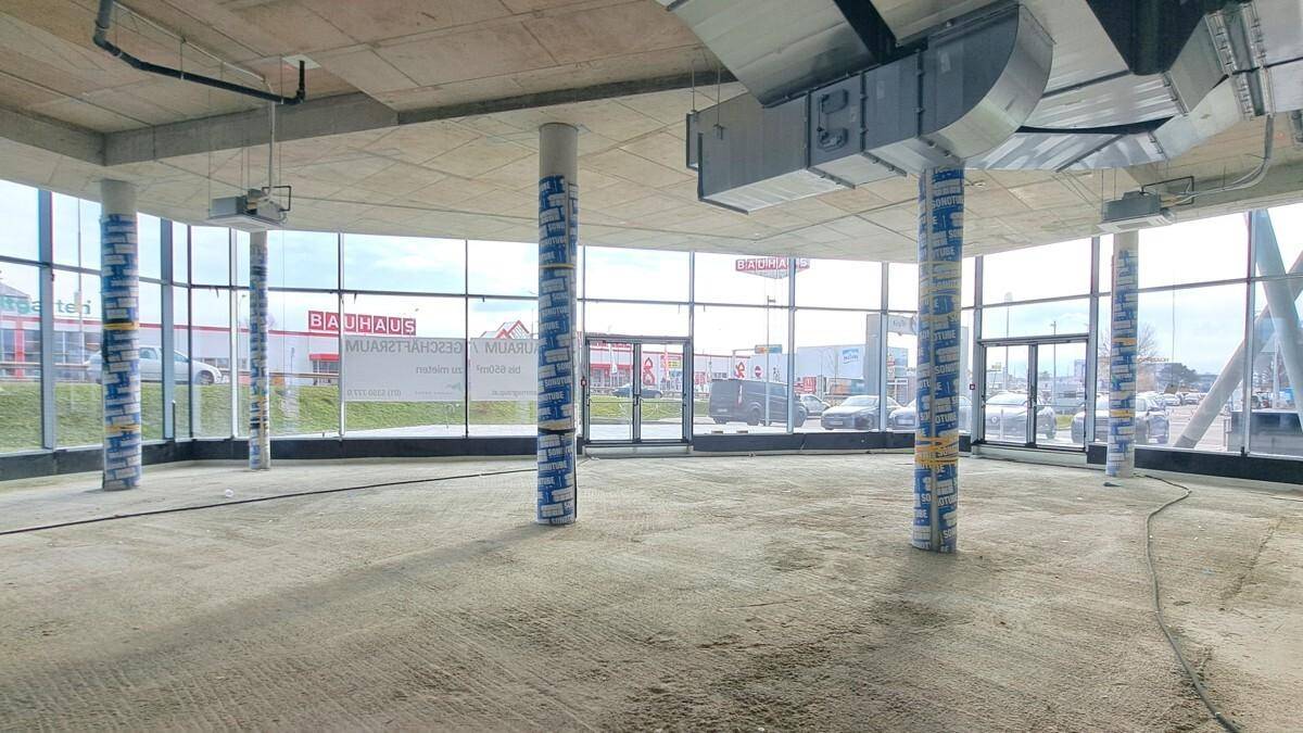 Großer, offener Gewerberaum im Rohbauzustand mit hohen Decken, sichtbaren Lüftungskanälen und umlaufenden Fenstern.