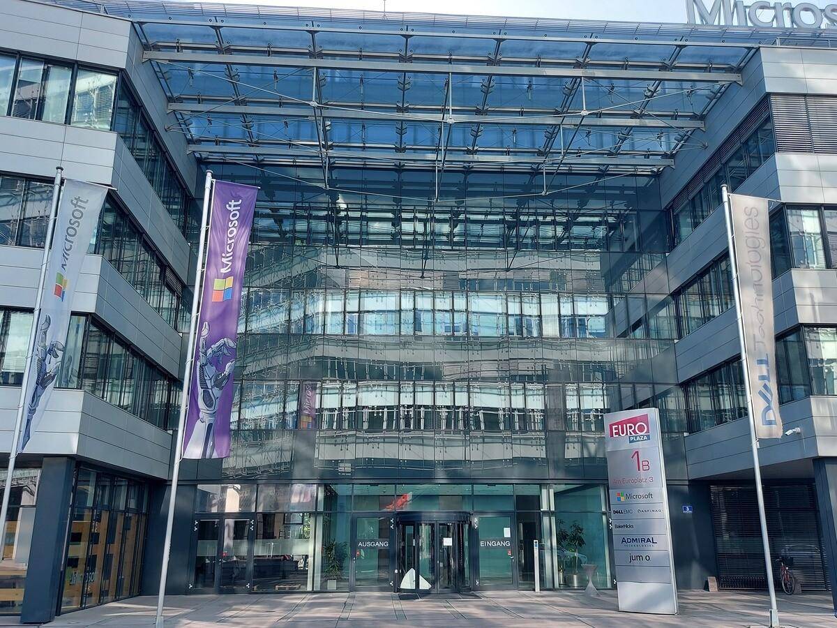 Eingangsbereich eines modernen Bürokomplexes mit Glasdach, Firmenflaggen und Hinweisschildern.