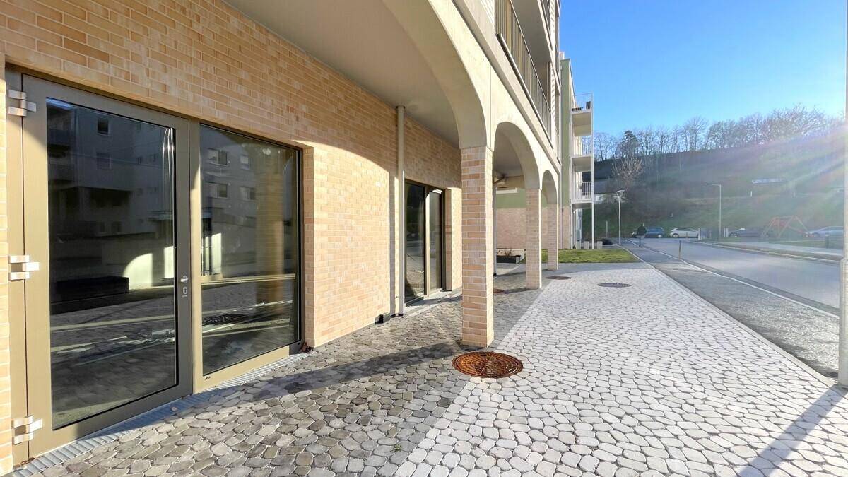 Eingangsbereich des Neubaus mit großen Glasfronten und gepflastertem Gehweg unter strahlendem Sonnenschein.