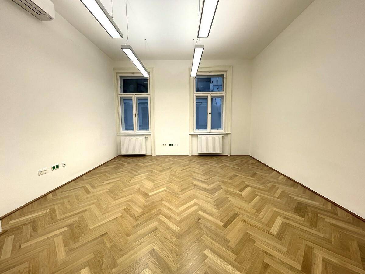 Heller Büroraum mit Fischgrätparkett und zwei großen Fenstern, die für eine angenehme Arbeitsatmosphäre sorgen.