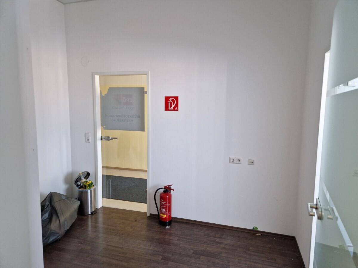 Leerer Innenraum mit weißen Wänden, dunklem Laminatboden, einer Glastür und einem Feuerlöscher.