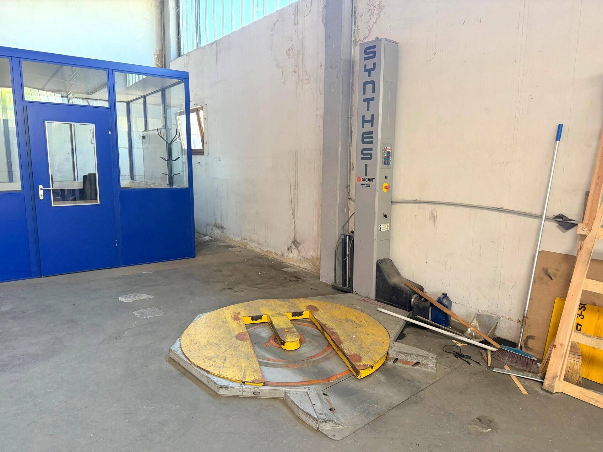 Industriebereich mit einer blauen Bürobox, einer Palettenwickelmaschine und einem Betonboden.