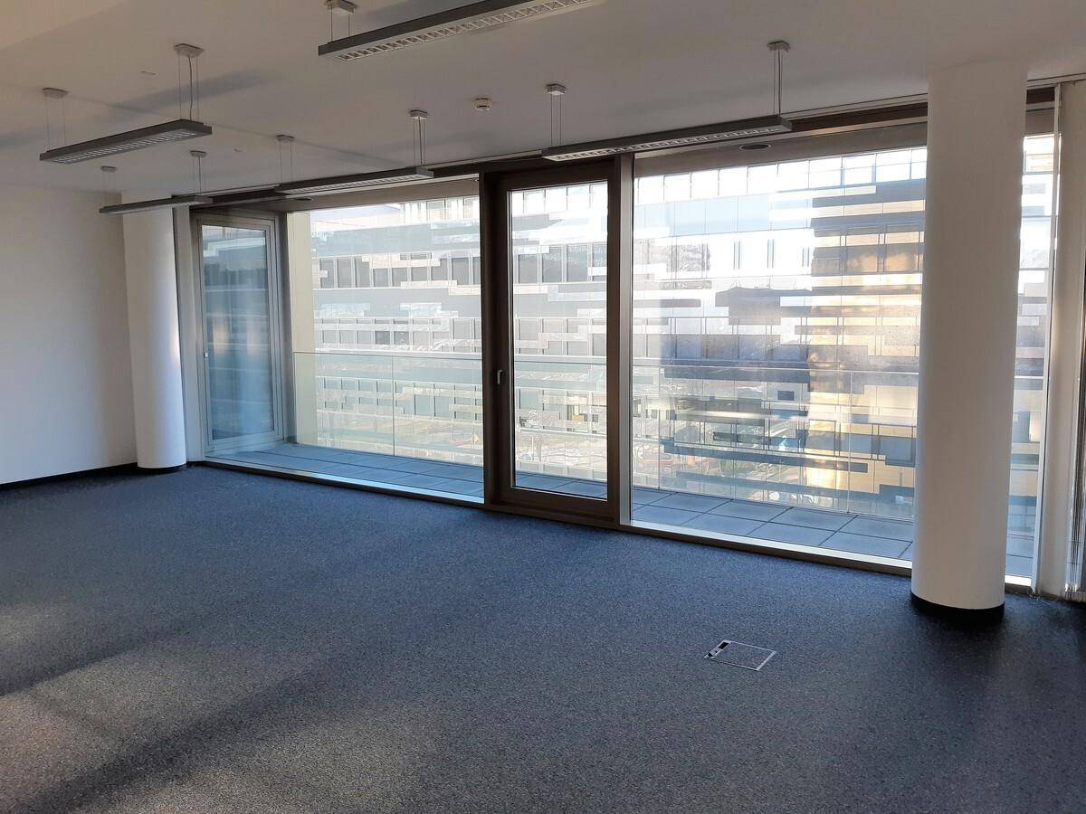 Leeres Büro mit dunklem Teppichboden und raumhohen Fenstern, die einen Blick auf die Stadt ermöglichen.