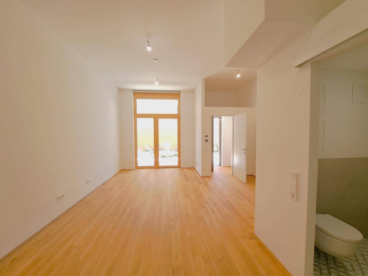 Heller, leerer Wohnraum mit Holzboden und großer Terrassentür, die viel Tageslicht hereinlässt.