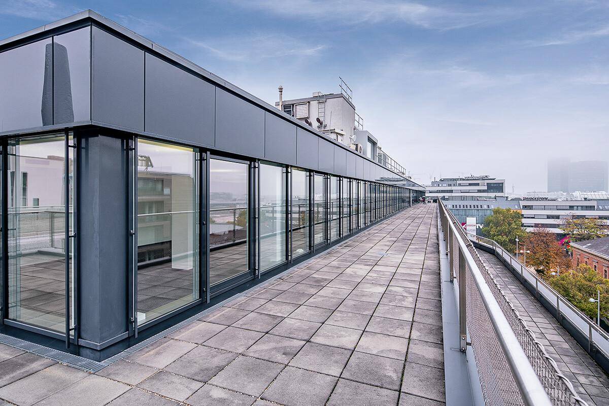 Lange Dachterrasse mit Fliesenboden und Glasgeländer, die einen weiten Blick über die umliegende Stadtlandschaft bietet.