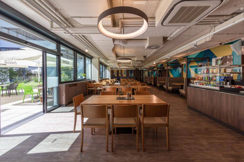 Helle Cafeteria mit Zugang zu einer Außenterrasse, Holztischen und einer modernen Essensausgabe.
