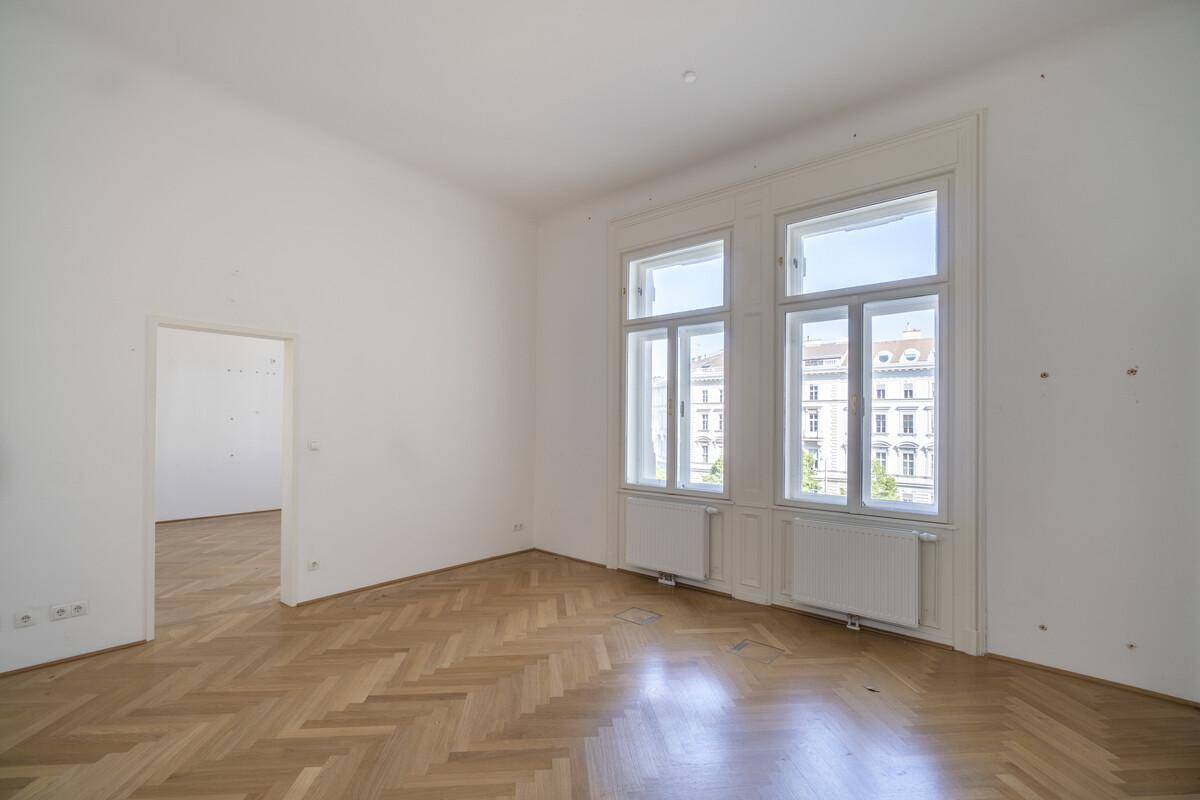 Geräumiger, heller Raum mit klassischem Fischgrätparkett und hohen Fenstern, die einen Blick nach draußen bieten.