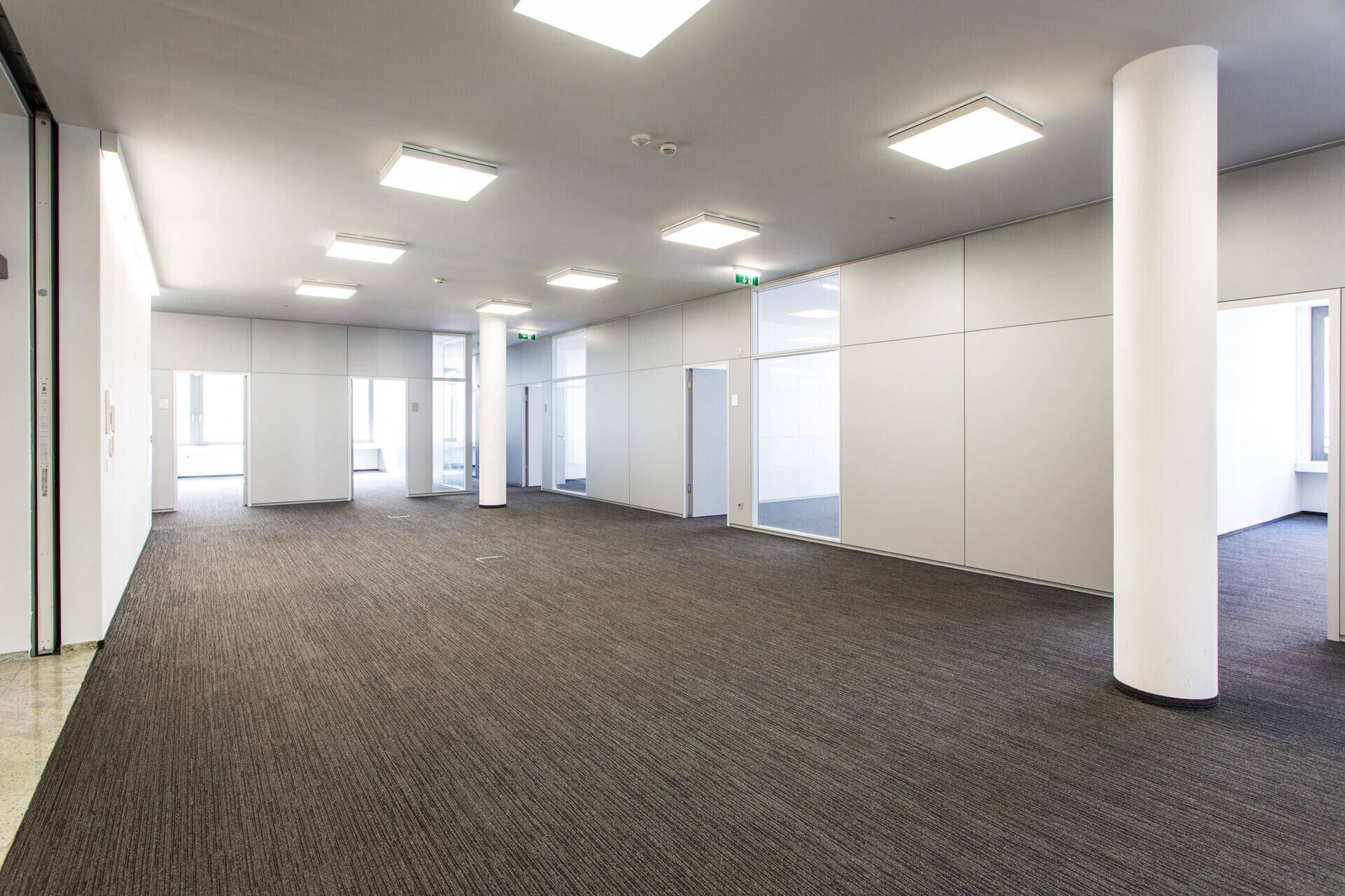 Großzügiger, offener Bürobereich mit Teppichboden, Säulen und modernen Deckenleuchten.
