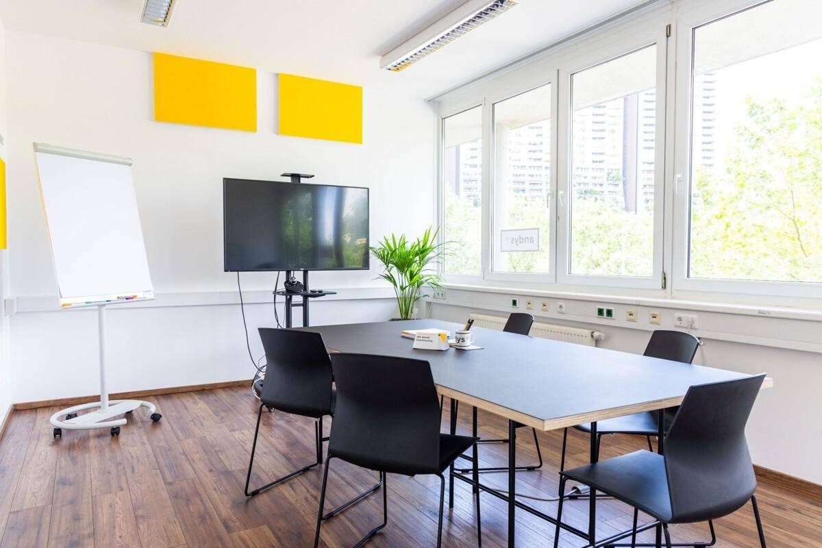Moderner Konferenzraum mit großem Tisch, Bildschirm und Flipchart, ideal für Besprechungen und Präsentationen.