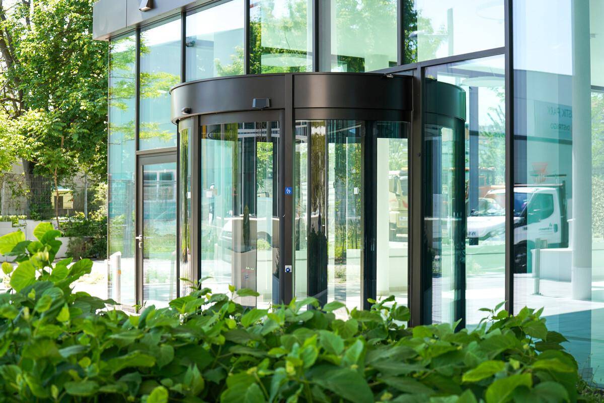 Moderner Eingangsbereich eines Bürogebäudes mit einer großen Drehtür aus Glas und umgebender Bepflanzung.