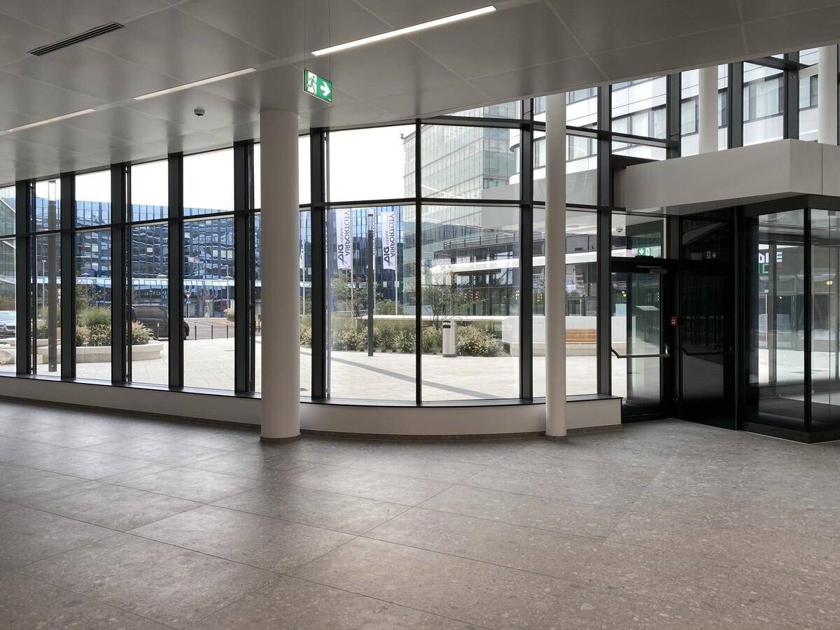 Heller Eingangsbereich eines modernen Gebäudes mit großen Glasfenstern und grauem Fliesenboden.