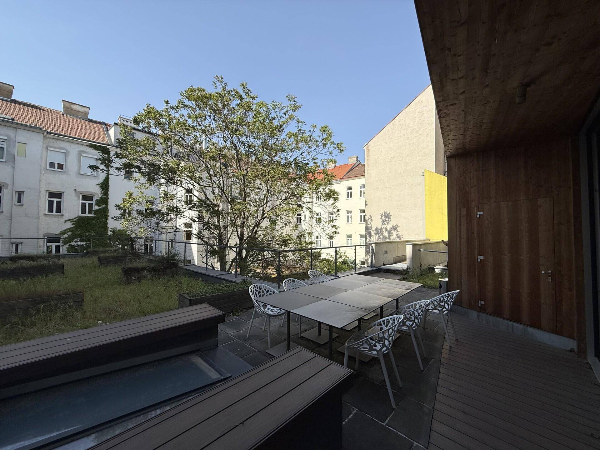 Große Außenterrasse mit Holzboden, Steinfliesen und Sitzgelegenheiten, umgeben von Bäumen.
