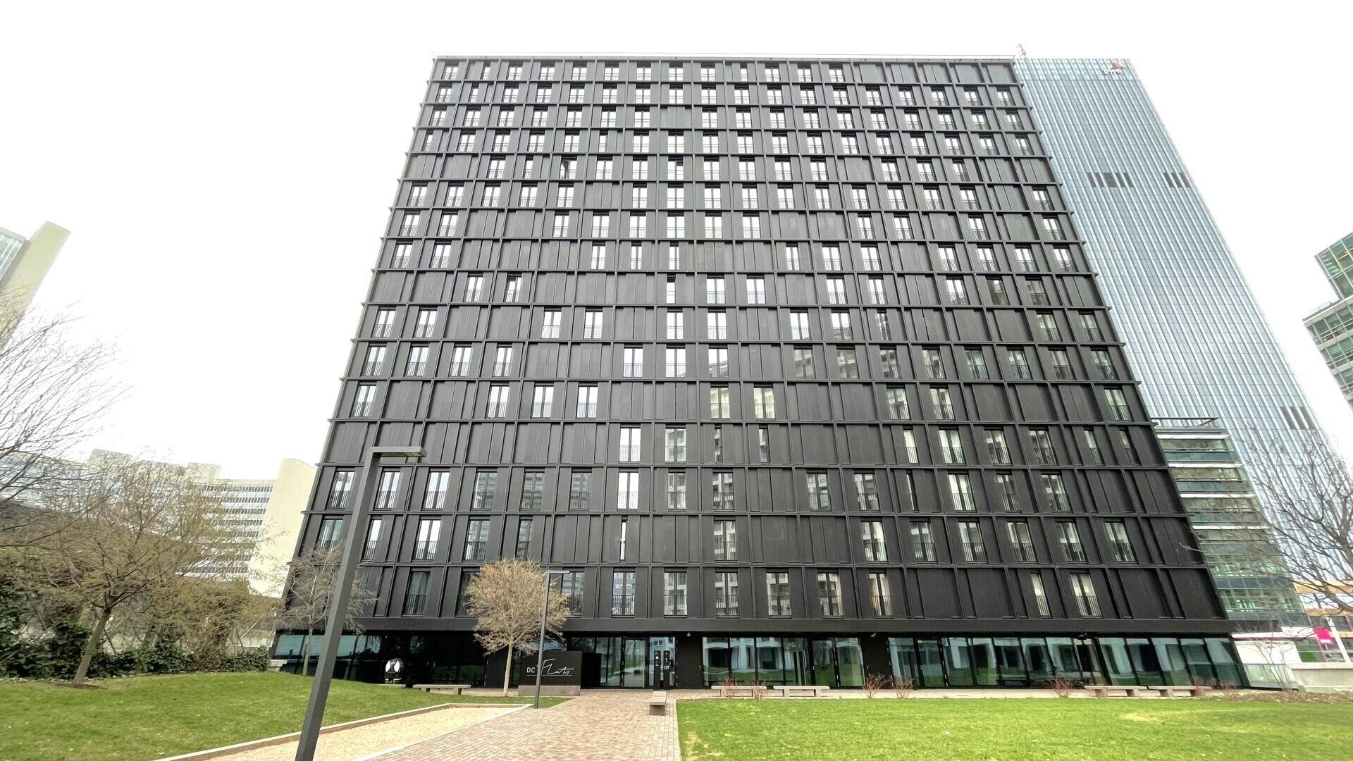 Gesamtansicht des modernen, dunklen Hochhauses mit vielen Fenstern und einer weitläufigen Grünfläche im Vordergrund.