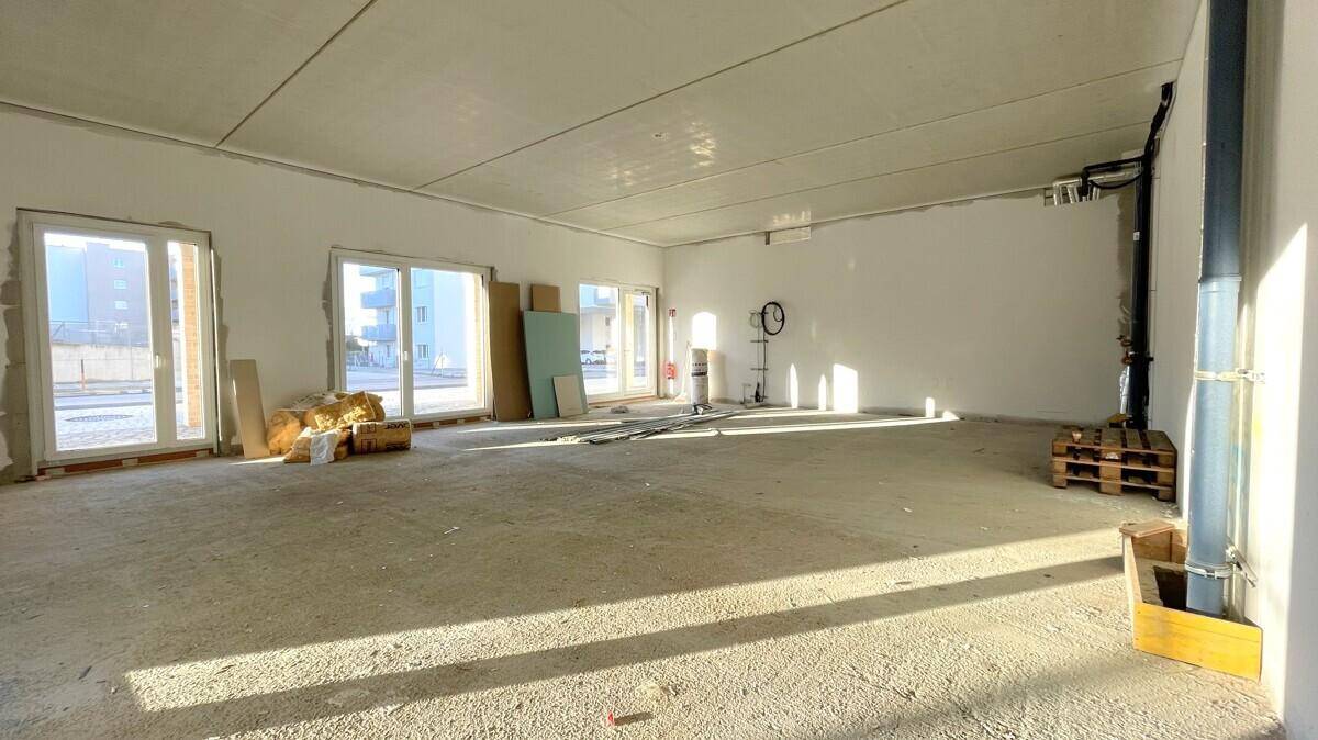 Rohbau-Zustand eines großen Raumes mit sichtbaren Versorgungsleitungen und viel natürlichem Licht.