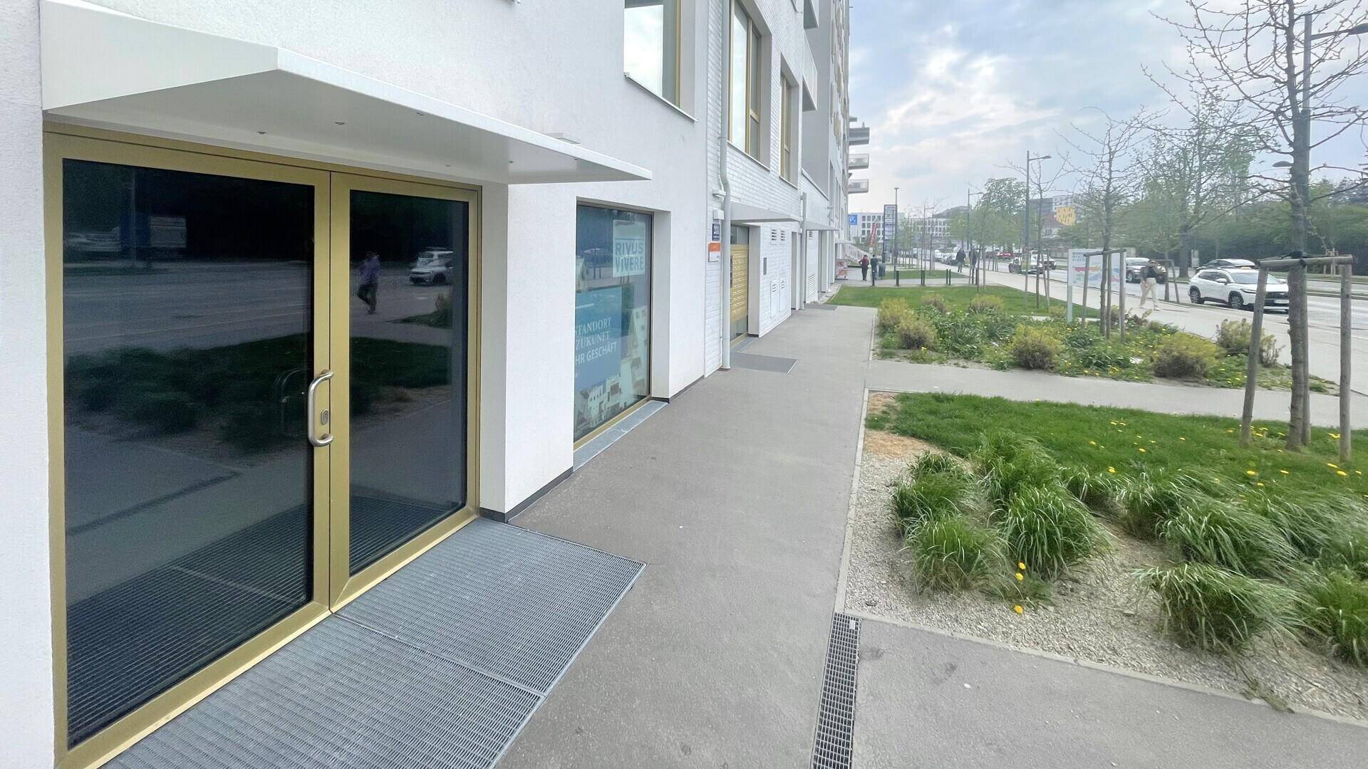 Eingangsbereich eines modernen Geschäftsgebäudes mit breitem Gehweg und Grünflächen entlang der Straße.
