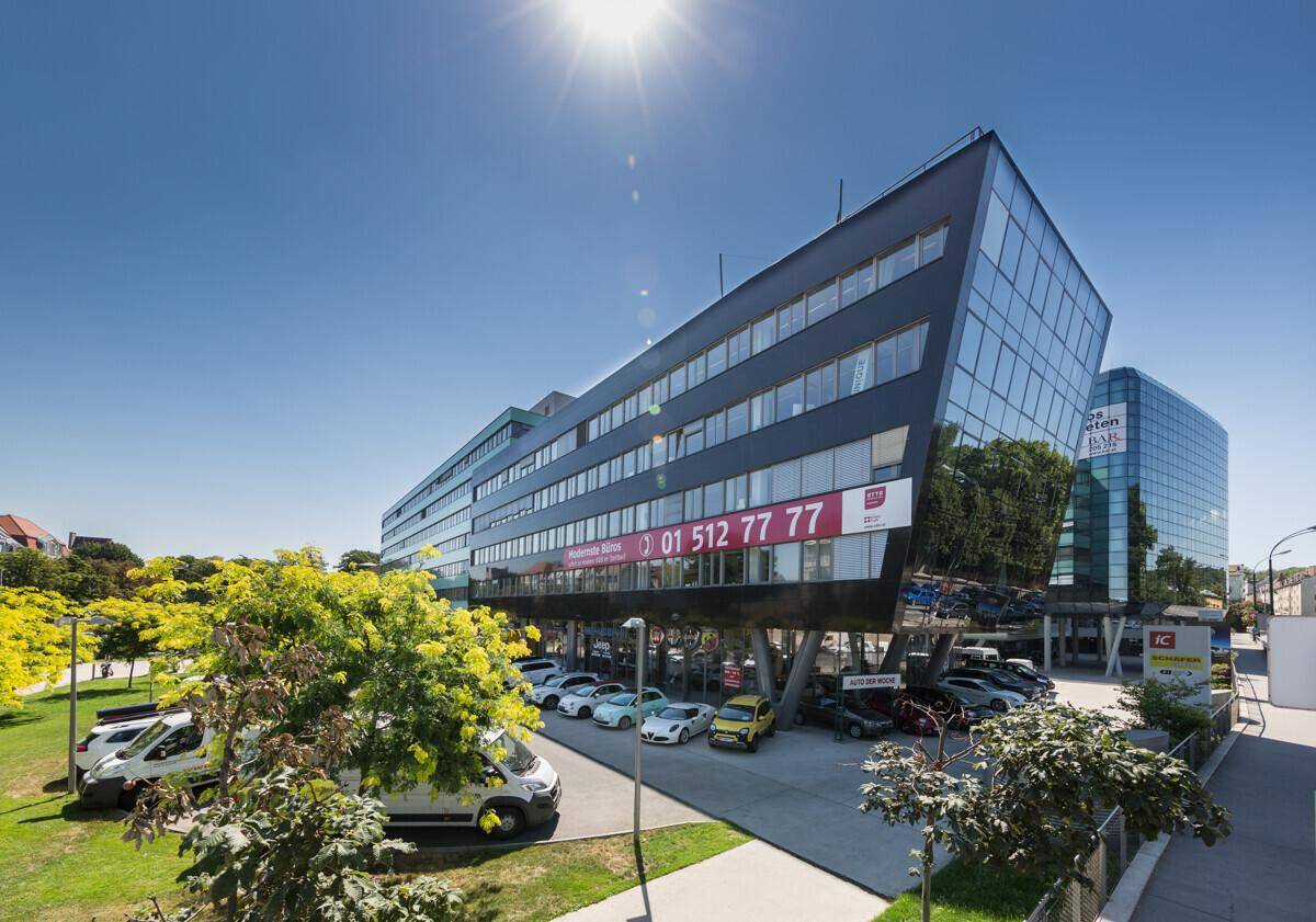 Ansicht des modernen Bürokomplexes mit auffälliger Glasfassade und davorliegendem Parkplatz unter strahlendem Himmel.