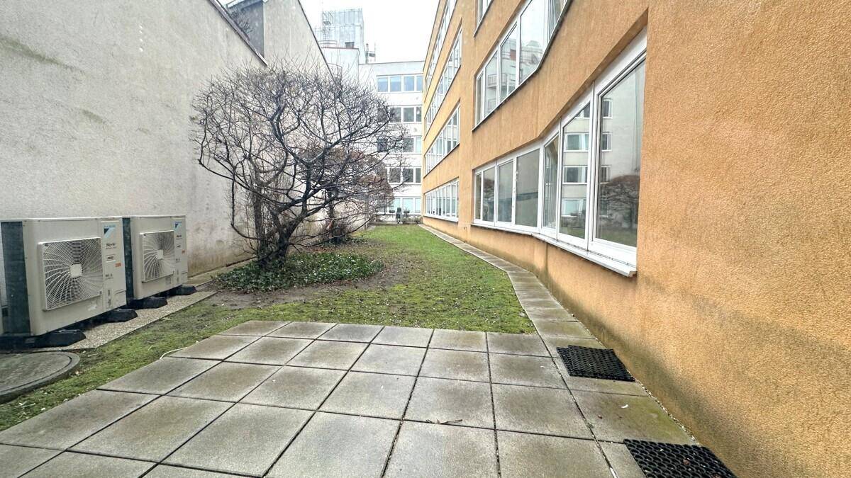 Außenbereich mit gepflasterter Terrasse, Rasenfläche und Klimaanlagen-Außengeräten an der Gebäudewand.