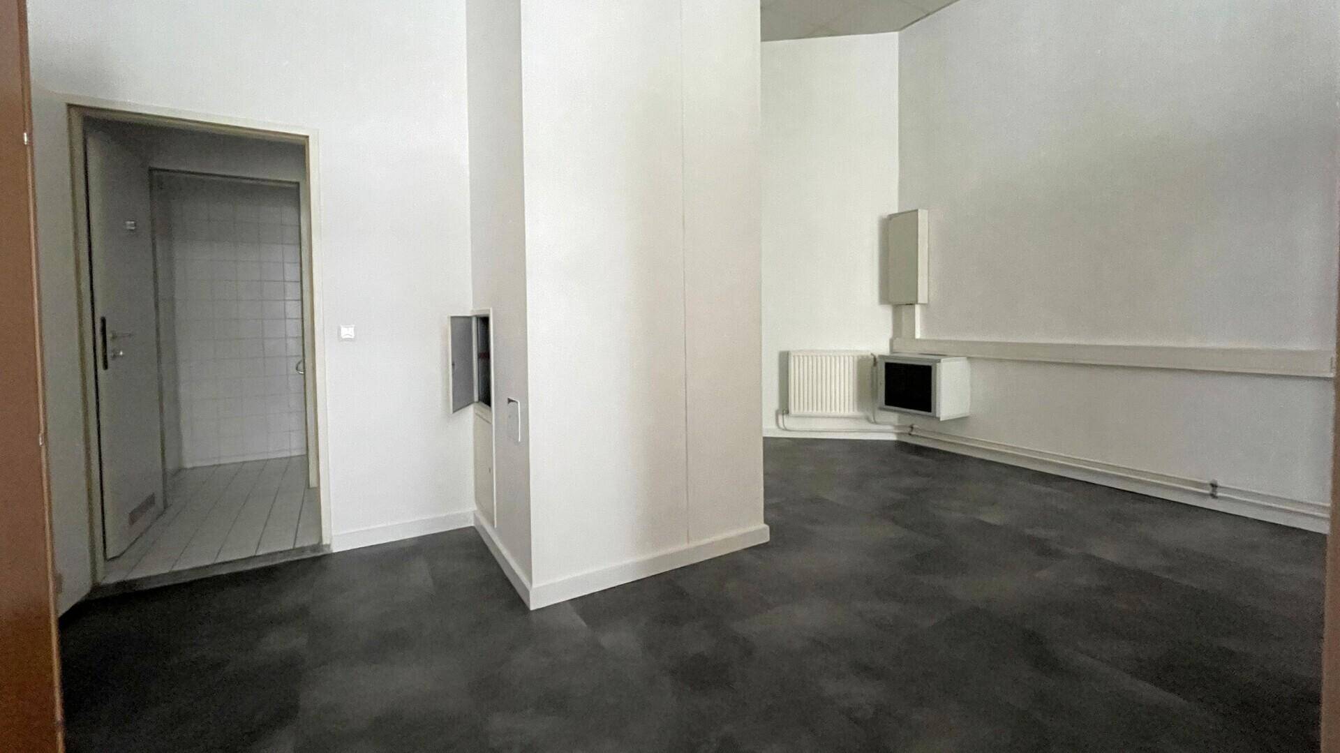 Büroraum mit dunklem Boden, weißem Pfeiler und Blick in einen gefliesten Nebenraum durch eine offene Tür.