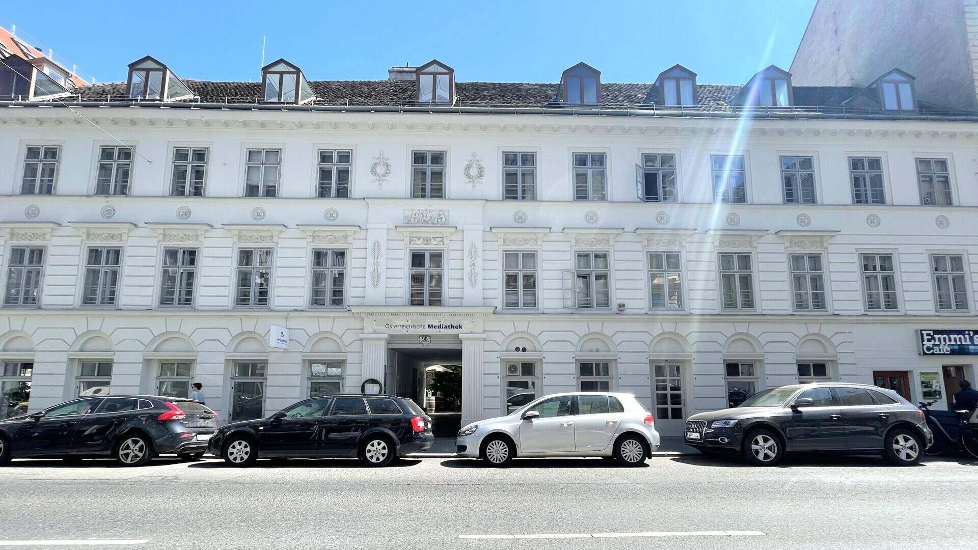 Klassisches weißes Gebäude mit vielen Fenstern und geparkten Autos auf der Straße davor unter blauem Himmel.