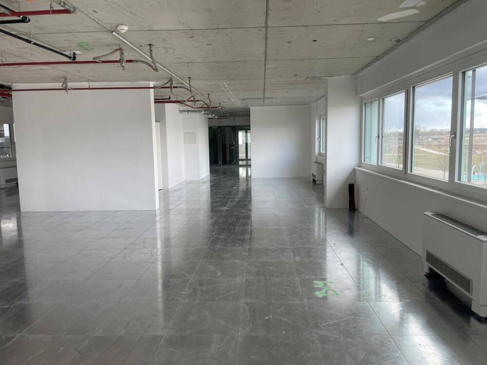 Großer, leerer Gewerberaum mit Fliesenboden, sichtbaren Leitungen an der Decke und großen Fenstern.