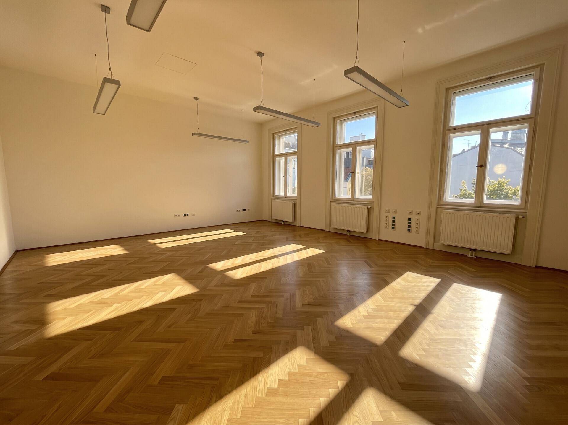 Heller Büroraum mit Fischgrätparkettboden, großen Fenstern und viel natürlichem Tageslicht, ideal für Arbeit.