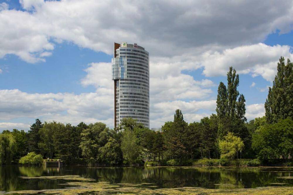 Hohes Bürogebäude am Seeufer, umgeben von üppiger grüner Vegetation unter einem bewölkten Himmel.