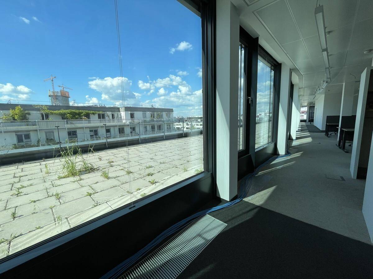 Langer Flur in einem Bürogebäude mit großen Fenstern und Blick auf eine begrünte Dachterrasse.