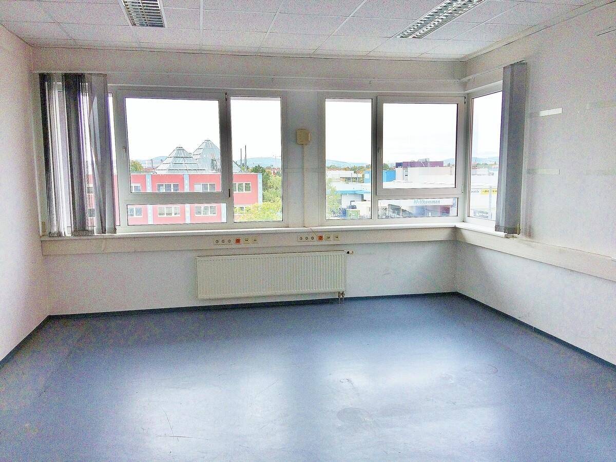 Heller Büroraum mit blauem Vinylboden, weißen Wänden und mehreren Fenstern, die viel Licht spenden.