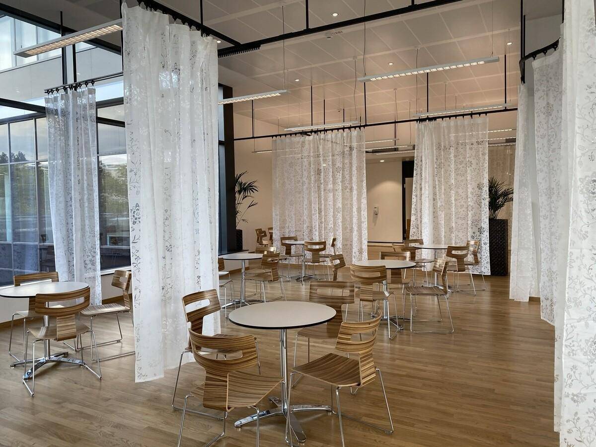 Helle Cafeteria mit Holztischen und -stühlen, umgeben von transparenten Vorhängen und Holzboden.