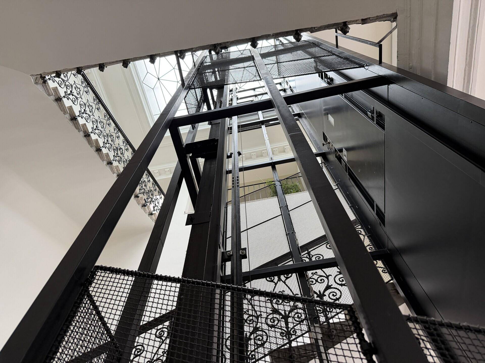 Moderner Aufzugsschacht aus Metall und Glas in einem historischen Treppenhaus mit Oberlicht.