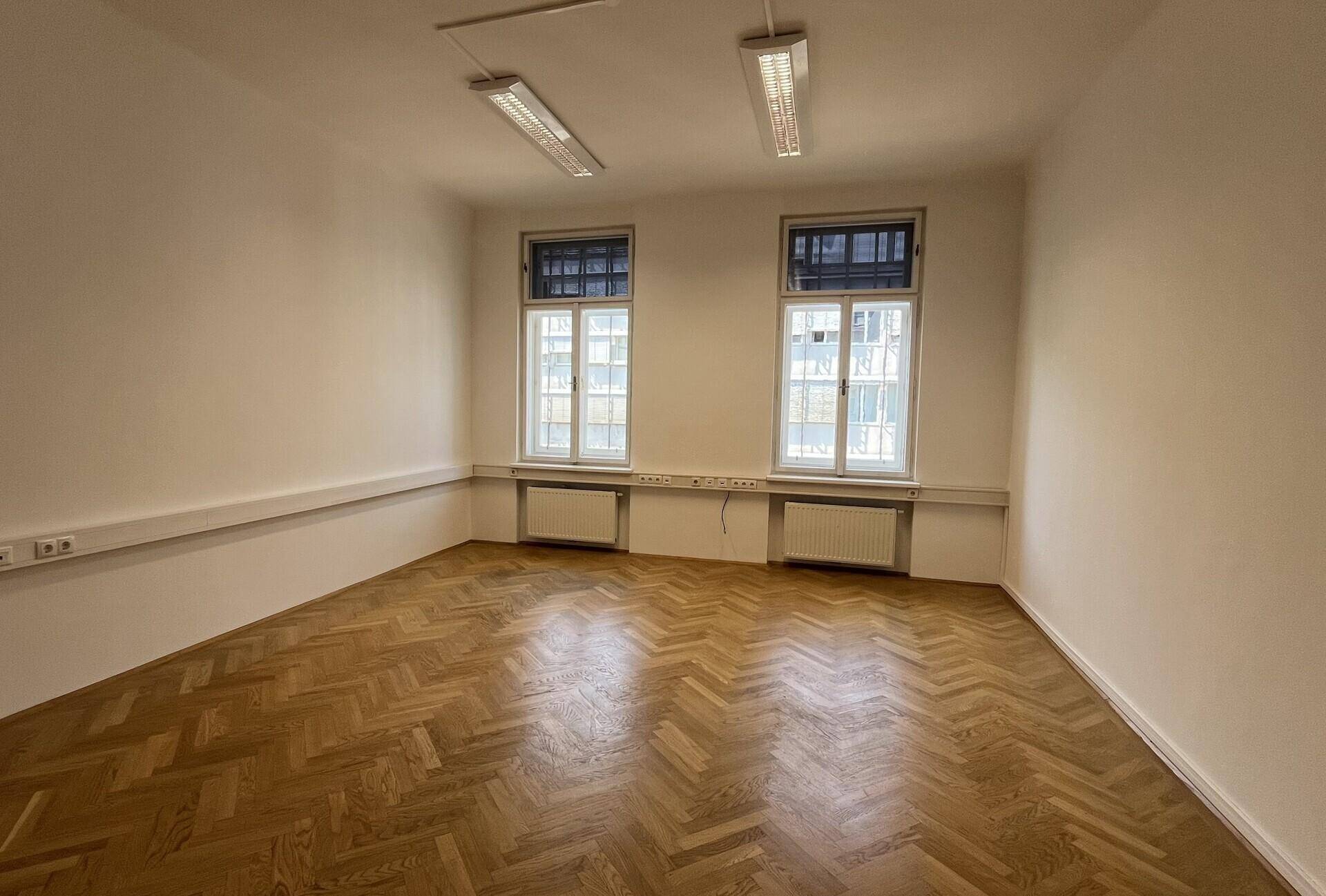 Leeres Büro mit Fischgrätparkett, hohen Fenstern und Wandheizkörpern, gut beleuchtet.