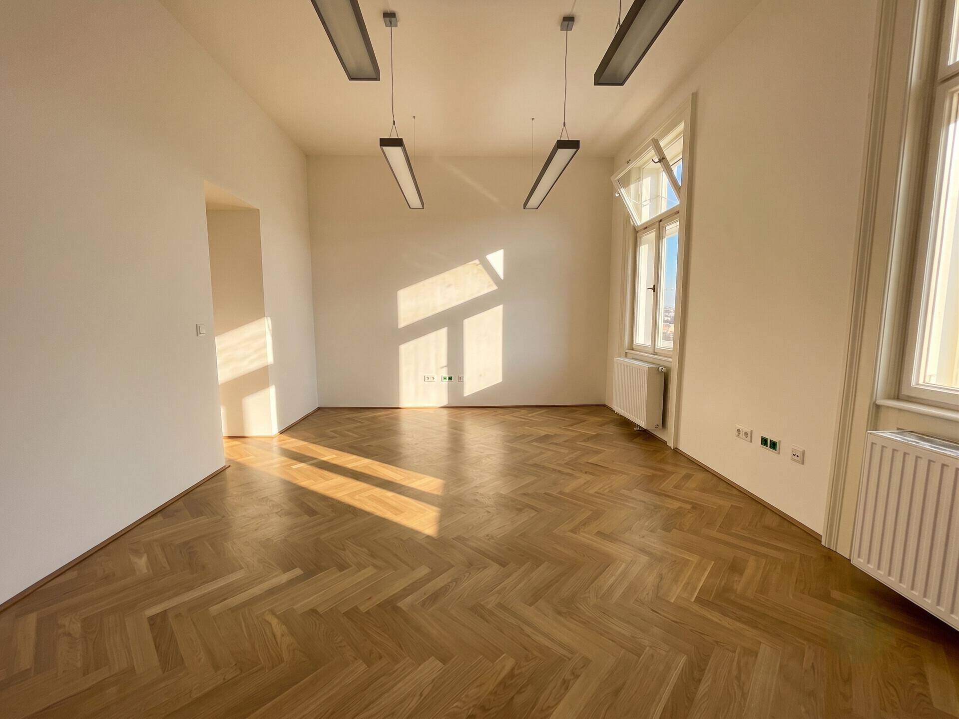Geräumiger Büroraum mit Fischgrätparkett, hohen Decken und viel natürlichem Licht durch die Fenster.