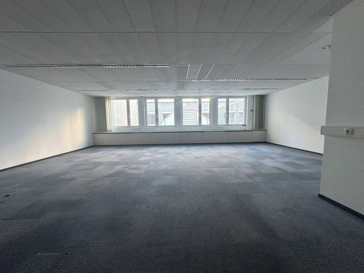 Weitläufiger Büroraum mit grauem Teppichboden und großen Fenstern, die für viel Helligkeit sorgen.
