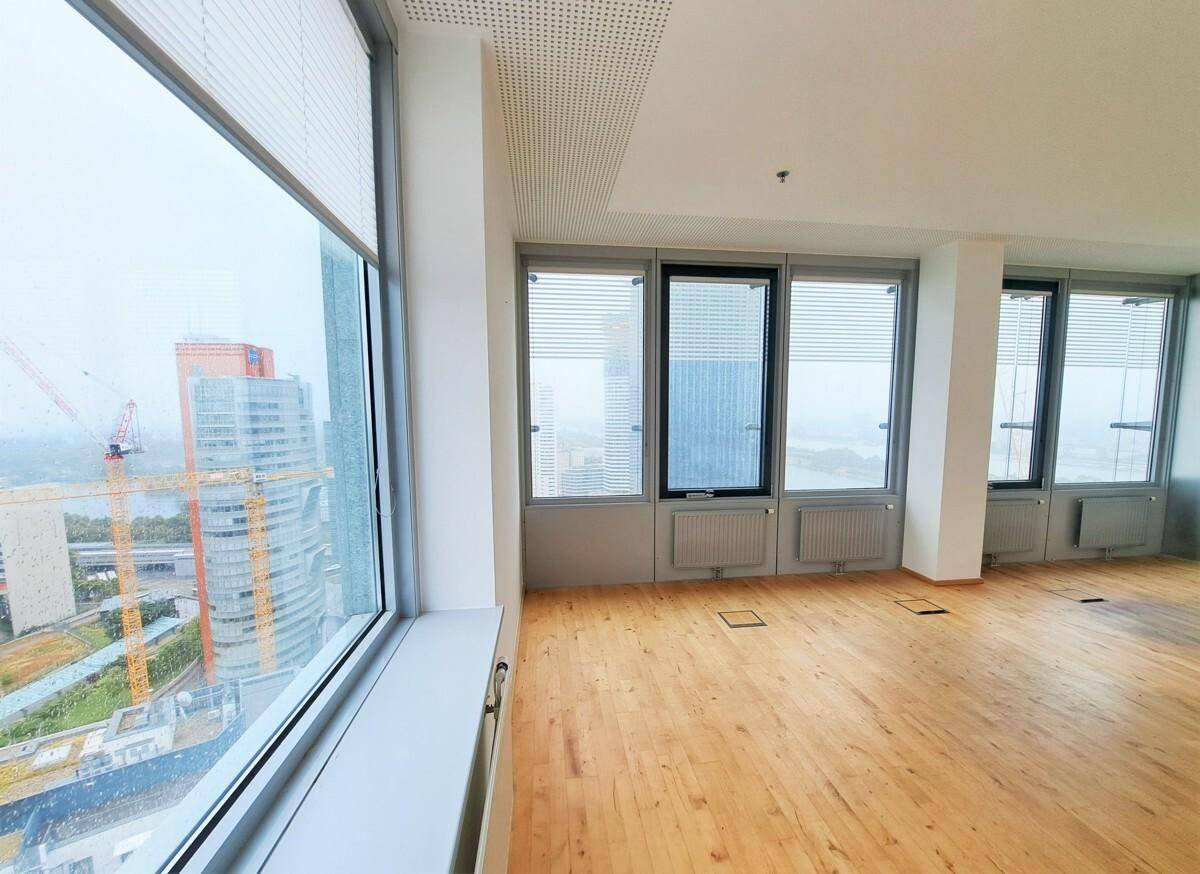Heller Raum mit Holzboden und Fenstern mit Jalousien, die einen Blick auf die Stadt und eine Baustelle ermöglichen.