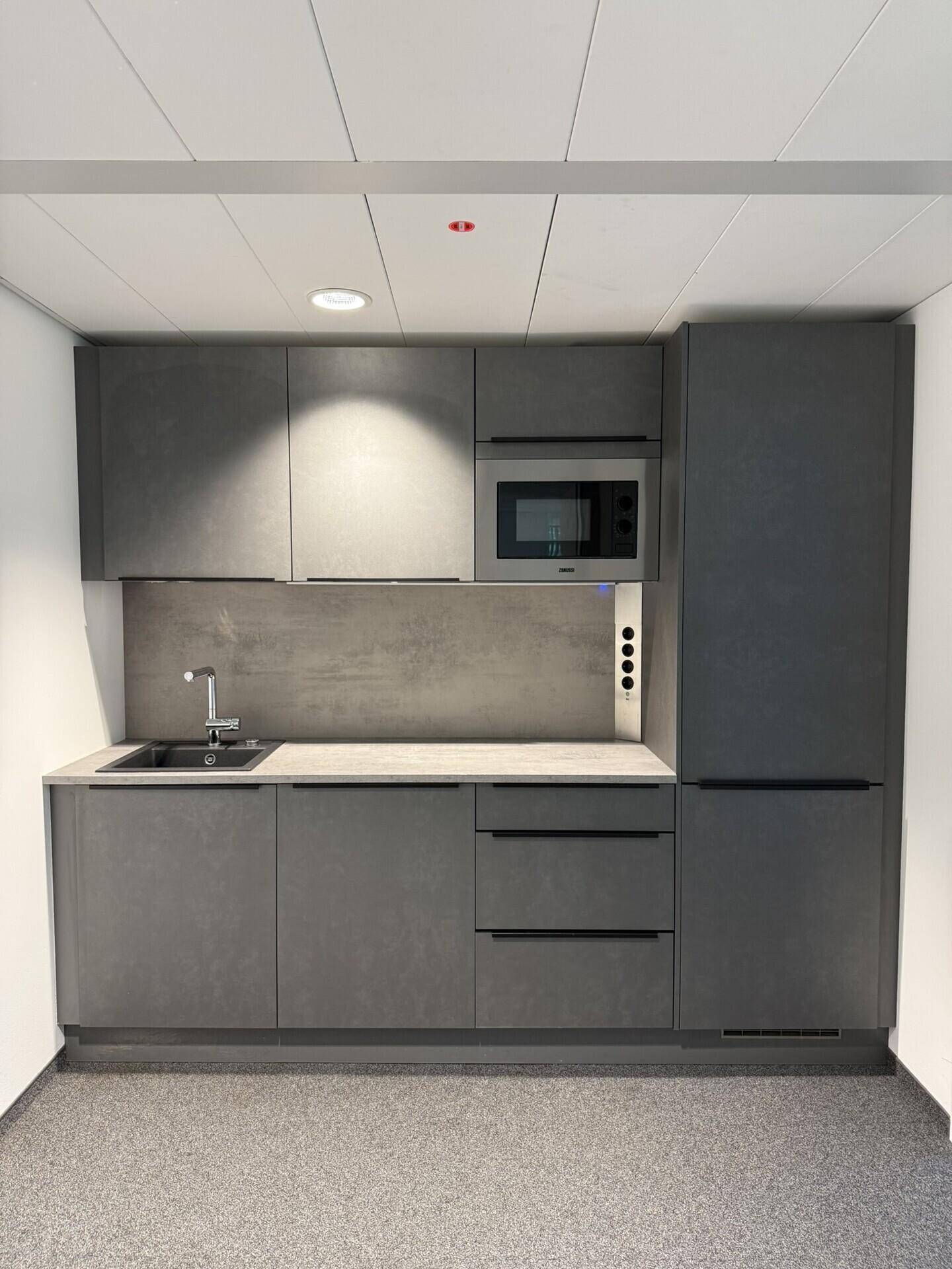 Moderne, kompakte Küchenzeile in Grau mit integrierter Mikrowelle und Kühlschrank, ideal für ein Büro.