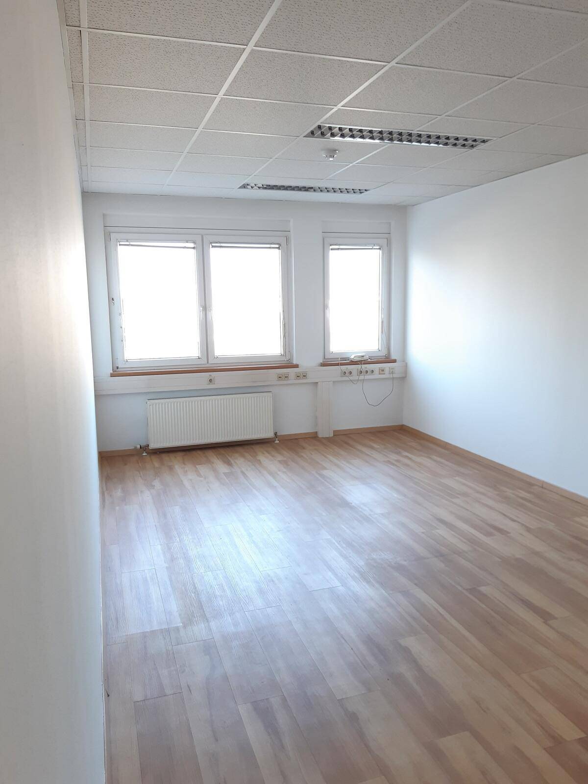 Leeres Bürozimmer mit hellem Laminatboden, weißen Wänden und einem Fenster mit Heizkörper.