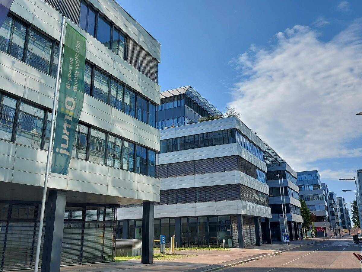 Reihe moderner Bürogebäude mit großen Fenstern und klaren Linien unter blauem Himmel, entlang einer Straße.