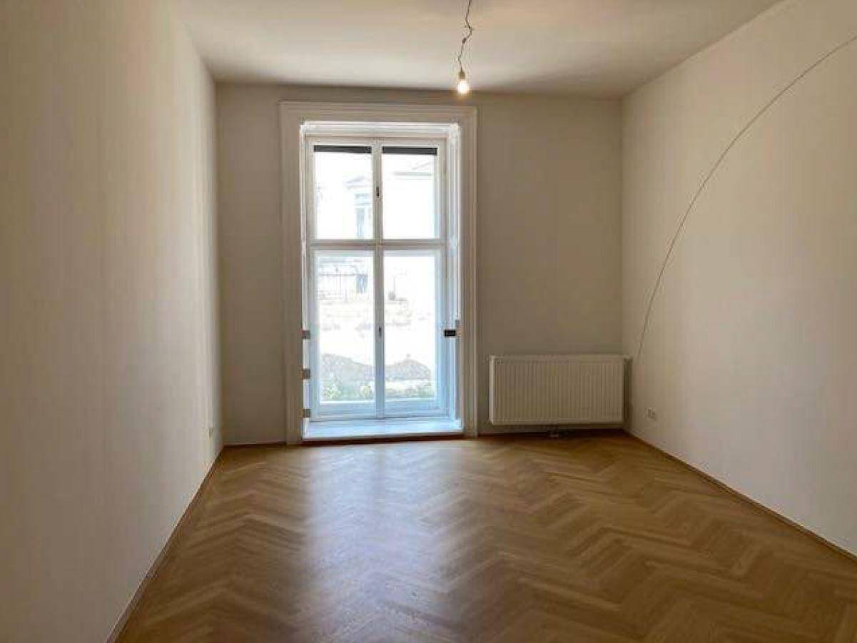 Helles Zimmer mit Fischgrätparkett, weiß gestrichenen Wänden und einem großen Fenster mit Heizkörper.