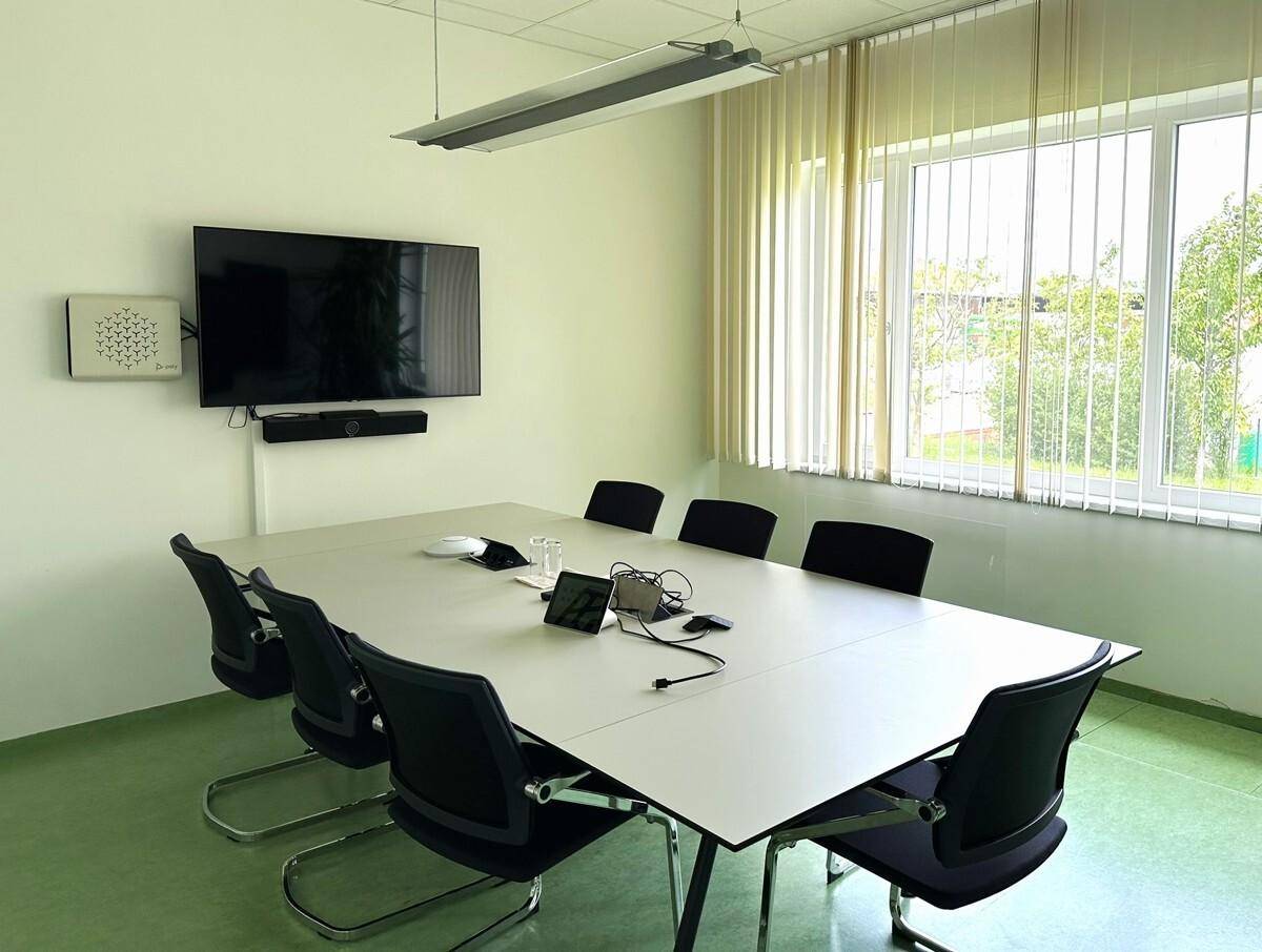 Kleinerer Besprechungsraum mit einem großen Bildschirm, einem Konferenztisch und Fenstern mit Jalousien.