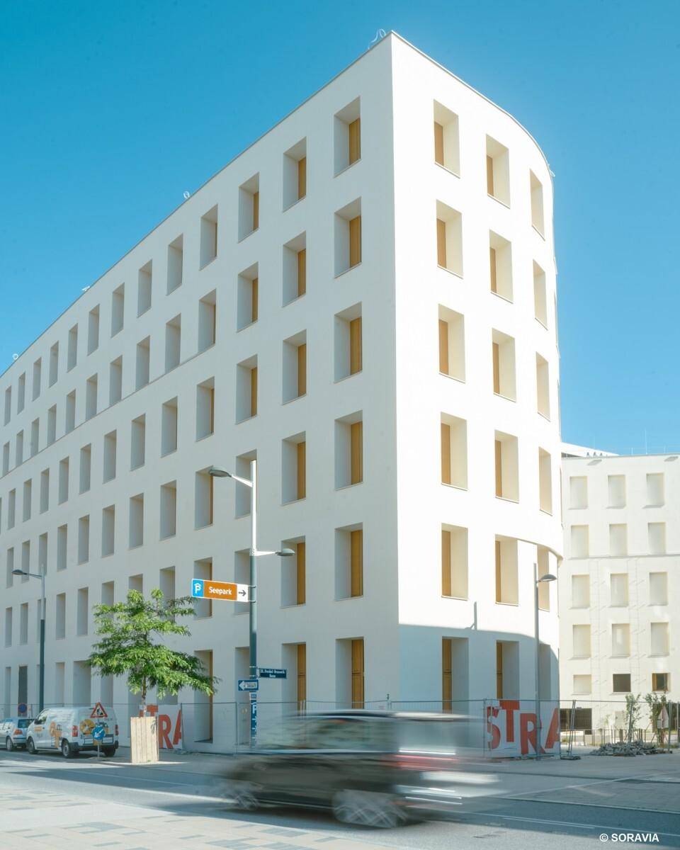 Moderne weiße Gebäudefassade an einer Straße mit Verkehr und einem Parkplatzschild.