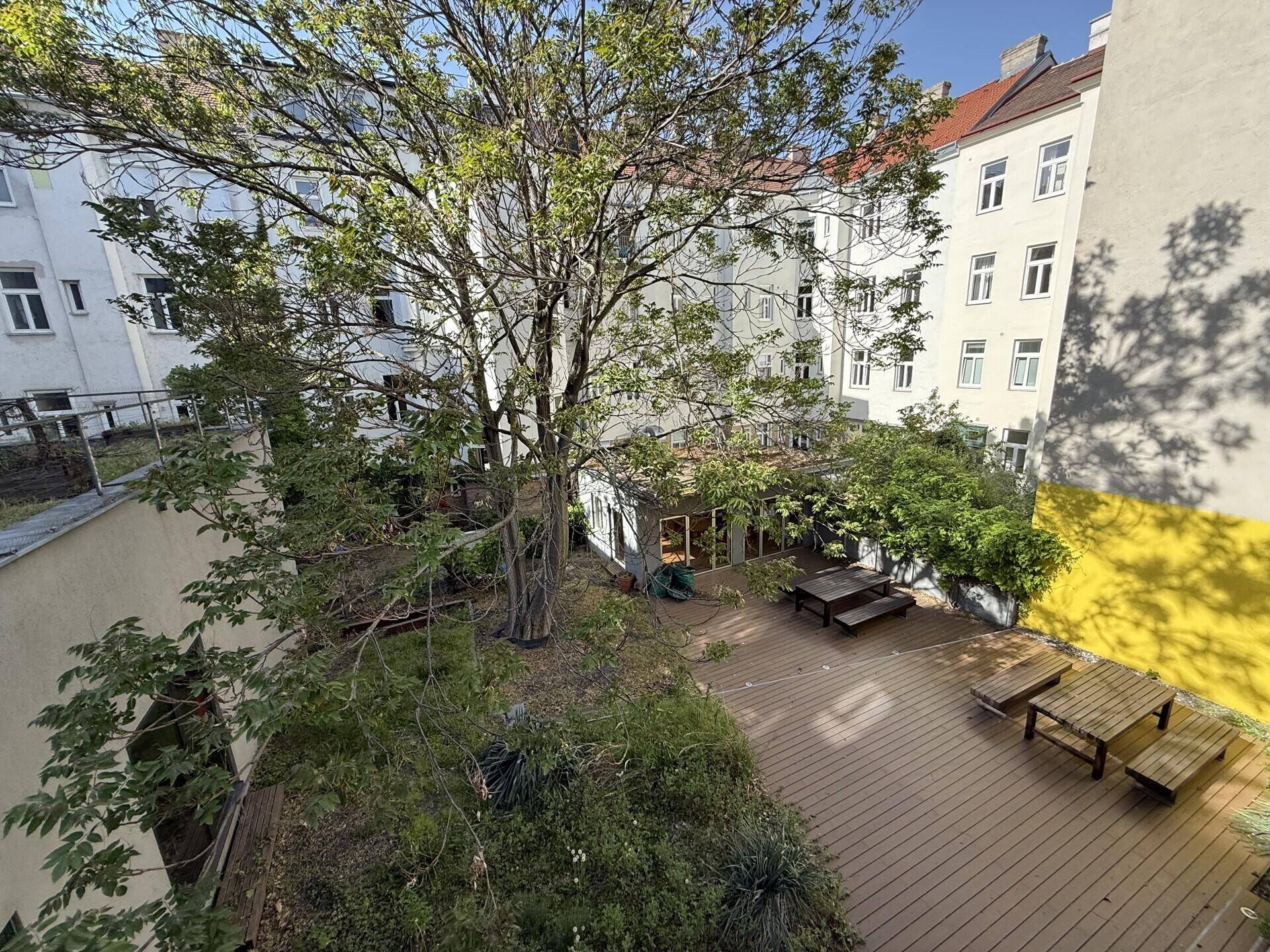 Begrünter Innenhof mit Holzboden, Sitzgelegenheiten und Bäumen, umgeben von Gebäuden.